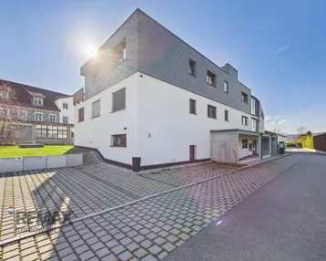 Modernes Mehrfamilienhaus mit weißer Fassade, Schieferdach und Außenparkplätzen unter strahlendem Himmel.