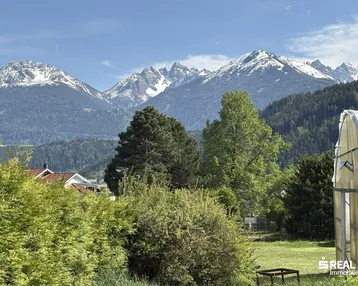 Panoramablick auf schneebedeckte Berge und grüne Landschaft mit vereinzelten Häusern.