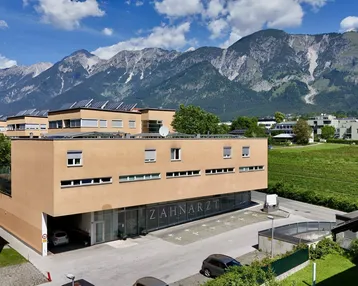 Gebäudeansicht mit Zahnarztpraxis, Parkplätzen und beeindruckendem Alpenpanorama im Hintergrund.
