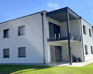 Modernes Mehrfamilienhaus mit weißer Fassade, Balkon und Gartenbereich unter blauem Himmel.