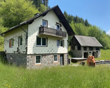 Haus mit Balkon und Nebengebäude in ländlicher Umgebung, umgeben von grünen Hügeln und Wald.