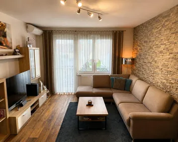 Gemütliches Wohnzimmer mit beiger Eckcouch, Holzboden und dekorativer Steinwand, hell und einladend.