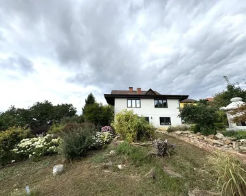 Weißes Haus auf einem Hügel mit üppiger Vegetation unter einem bewölkten Himmel.
