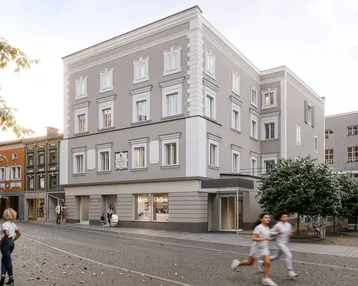 Graues Gebäude mit klassischer Fassade an einer belebten Straße mit Geschäften und Passanten.
