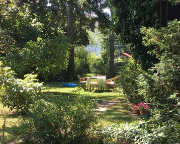 Idyllischer Gartenbereich mit Sitzgelegenheiten unter schattenspendenden Bäumen und blühenden Pflanzen.
