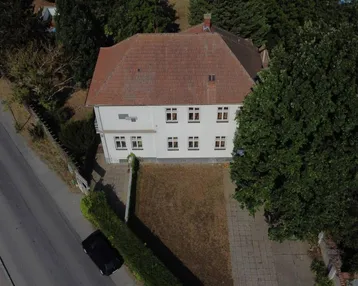 Luftaufnahme eines weißen Gebäudes mit rotem Ziegeldach, umgeben von Bäumen und einer Straße.