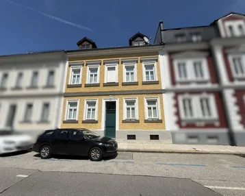 Gelbe Fassade eines Mehrfamilienhauses mit grüner Eingangstür und parkendem Auto auf der Straße.