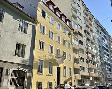 Gelbes Mehrfamilienhaus an einer sonnigen Straße mit parkenden Autos und angrenzenden Gebäuden.
