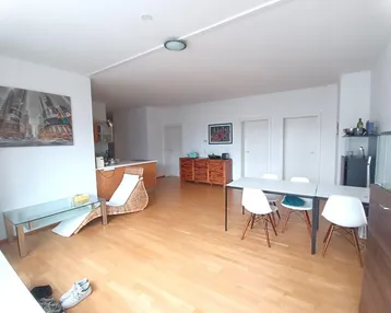 Helles, offenes Wohn-Esszimmer mit Holzboden und integrierter Küche im Hintergrund.