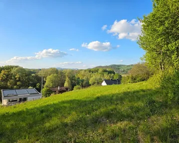 Grüne Hügellandschaft mit Bäumen und Häusern in der Ferne unter blauem Himmel.