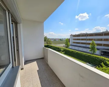 Heller Balkon mit Blick ins Grüne und auf die umliegende Bebauung unter blauem Himmel.