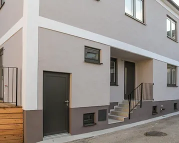 Außenansicht des Gebäudes mit moderner Fassade, Holzterrasse und Zugang zum Eingangsbereich.