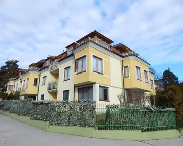 Mehrfamilienhaus mit gelber Fassade, roten Dächern und Balkonen unter bewölktem Himmel.