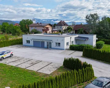 Luftaufnahme eines modernen Gebäudes mit großem Parkplatz und grüner Hecke, umgeben von ländlicher Landschaft.