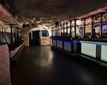 Dunkler Barbereich mit Höhlen-Design, hohen Tischen und blauer LED-Beleuchtung, ideal für Veranstaltungen.