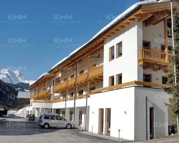 Modernes Gebäude im alpinen Stil mit Holzbalkonen und schneebedeckten Dächern, umgeben von Bergen.