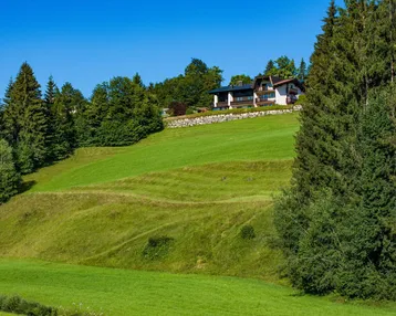 Haus in alpiner Landschaft mit grünen Wiesen und hohen Bäumen unter blauem Himmel.
