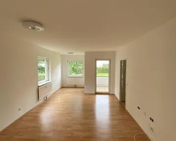 Geräumiger, leerer Wohnbereich mit hellem Laminatboden und Zugang zum Balkon.