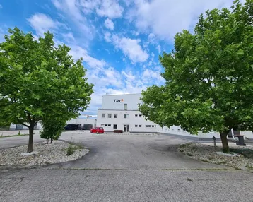 Weißes Geschäftsgebäude mit Firmenlogo, umgeben von Bäumen und einem Parkplatz unter blauem Himmel.