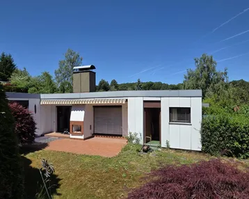 Modernes Haus mit Flachdach, Terrasse und Außenkamin, umgeben von viel Grün unter blauem Himmel.
