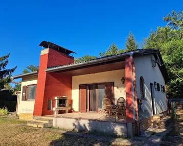 Hausfassade mit überdachter Terrasse, Außenkamin und Sitzgelegenheiten im Freien unter blauem Himmel.