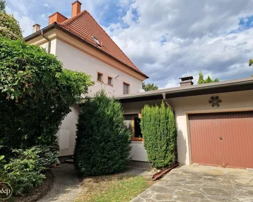 Hausfassade mit rotem Ziegeldach, angrenzender Garage und gepflegtem Vorgarten unter bewölktem Himmel.