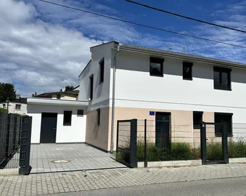 Moderne Hausfassade mit weißem und beigem Putz, dunklen Fenstern und gepflasterter Einfahrt.