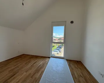 Leeres Zimmer mit Holzboden, schrägen Wänden und einem hohen Fenster mit Blick auf die Umgebung.