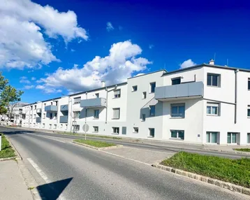 Moderne Wohnanlage mit weißer Fassade und grauen Balkonen entlang einer Straße unter blauem Himmel.