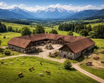 Luftaufnahme eines weitläufigen Anwesens mit mehreren Gebäuden, umgeben von grünen Wiesen und majestätischen Bergen im Hintergrund.