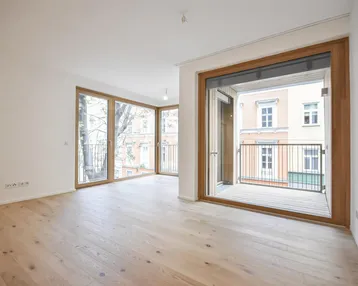 Helles Wohnzimmer mit Holzboden und Zugang zu einem kleinen Balkon, umgeben von Stadtbäumen.