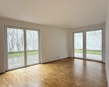 Helles Zimmer mit Holzboden und großen Fenstertüren, die ins Grüne führen.