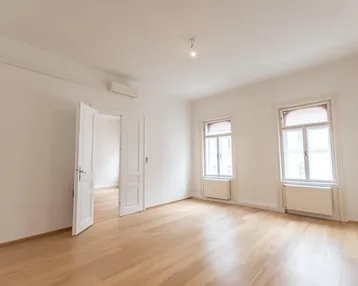 Helles, leeres Zimmer mit Parkettboden und zwei Fenstern, bereit zur individuellen Gestaltung.