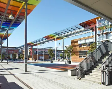 Weitläufiger Außenbereich eines modernen Gebäudekomplexes mit bunten Überdachungen, Treppen und einem Aufzug.