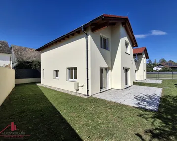 Neubau-Doppelhaushälfte mit kleiner Rasenfläche und gepflasterter Terrasse unter blauem Himmel.