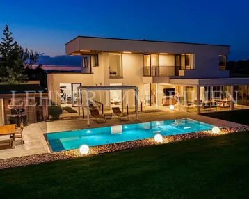 Moderne Villa mit beleuchtetem Pool, Garten und gemütlichem Essbereich im Freien bei Dämmerung.