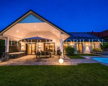 Elegantes Haus bei Nacht mit beleuchteter Terrasse, Pool und Whirlpool, umgeben von Grün.