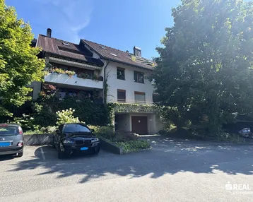Mehrfamilienhaus mit Parkplätzen und üppiger Begrünung unter blauem Himmel.