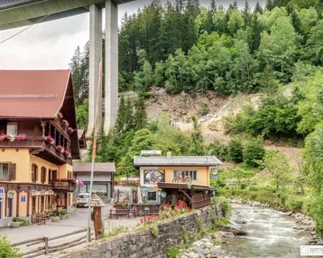 Traditionelle Gebäude am Flussufer mit einer Brücke im Hintergrund und bewaldeten Bergen.