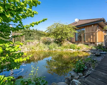 Außenansicht des Hauses mit Holzfassade, umgeben von einem naturnahen Garten mit Teich und üppiger Vegetation.