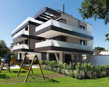 Modernes Mehrfamilienhaus mit gestuften Balkonen, Spielplatz und gepflegter Grünfläche unter blauem Himmel.