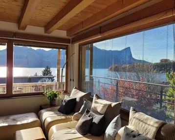 Helles Wohnzimmer mit Panoramablick auf den See und die umliegende Berglandschaft durch große Fenster.