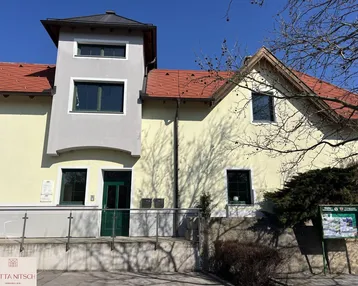 Gelbe Fassade eines Gebäudes mit grauem Anbau und rotem Ziegeldach unter blauem Himmel.
