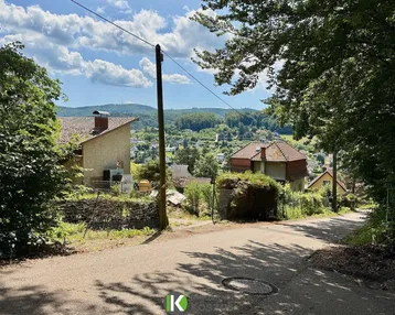 Panoramablick über eine hügelige Landschaft mit Häusern und viel Grün unter blauem Himmel.