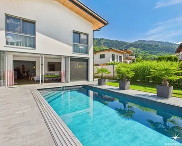 Modernes Haus mit Pool und Terrasse, umgeben von grüner Landschaft und Bergen.