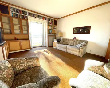 Helles Wohnzimmer mit Teppichboden, Bücherregalen und Zugang zum Balkon.