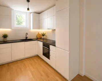 Moderne Einbauküche mit weißen Schränken, dunkler Arbeitsplatte und hellem Holzfußboden, gut beleuchtet.