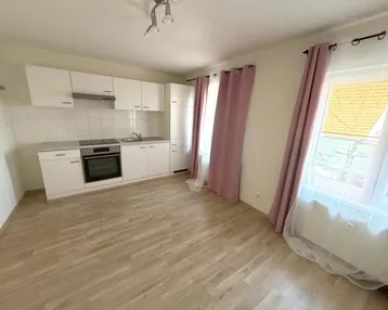 Helle Wohnküche mit weißer Einbauküche, Laminatboden und zwei Fenstern mit rosa Vorhängen.
