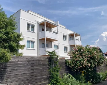 Modernes Mehrfamilienhaus mit weißer Fassade und mehreren Balkonen unter blauem Himmel.