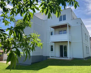 Modernes Mehrfamilienhaus mit weißer Fassade, Balkonen und umliegender Grünfläche unter blauem Himmel.